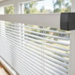A bright home window scene, a matte dark-gray 3D printed end cap adapter fitted neatly to the end of a white blind headrail