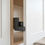 A bright kitchen scene, a white cabinet door held closed by a matte charcoal-gray 3D printed catch plate fitted behind a small magnetic catch inside