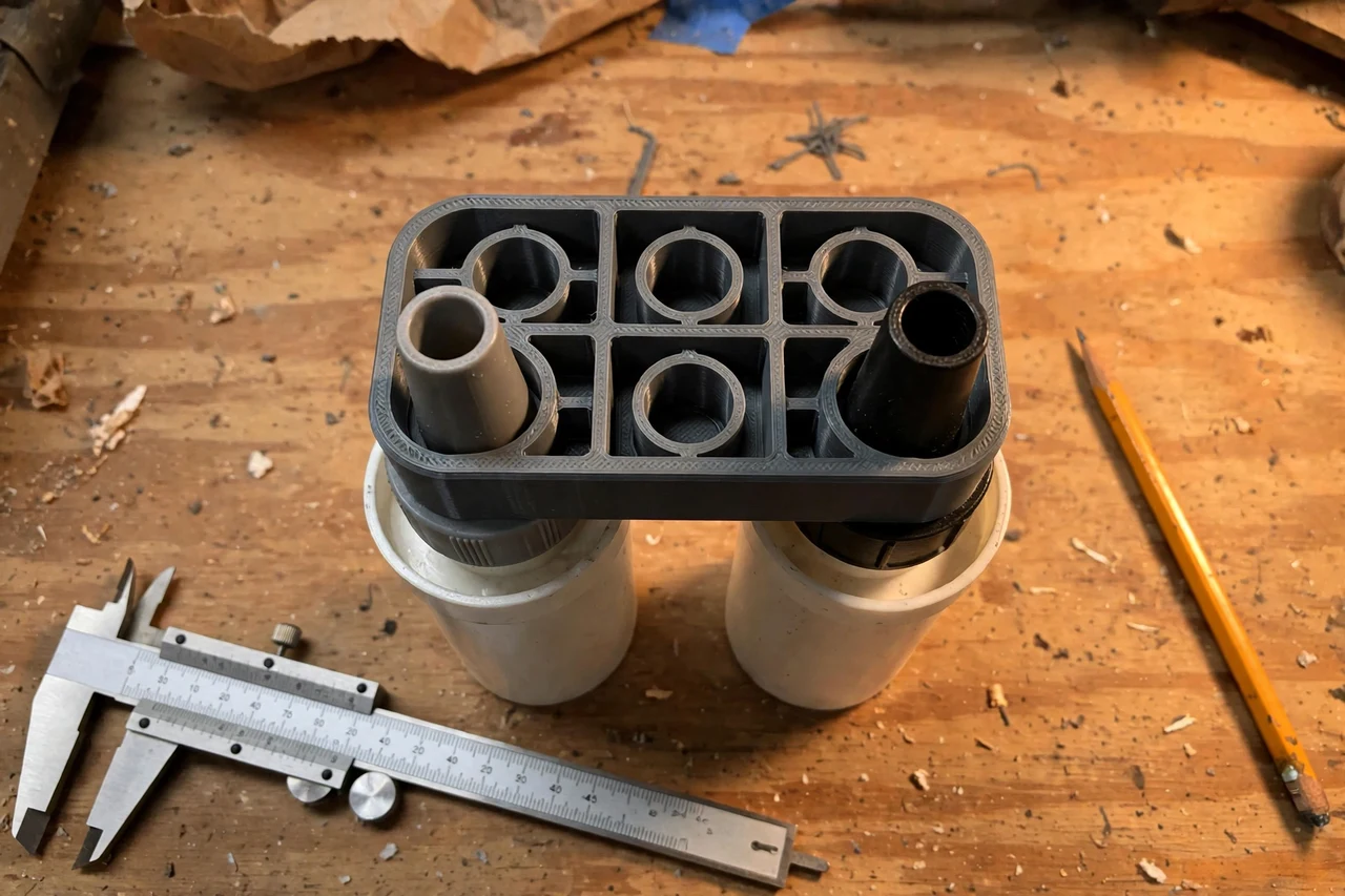 Close-up top-down view of a matte charcoal-gray 3D printed caulk gun nozzle cap holders being test-fitted against caulk tube nozzles capped between uses on a workbench, calipers and a pencil nearby, warm workshop lighting