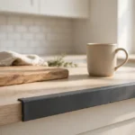 A bright kitchen scene, a laminate countertop edge fitted with a neat matte charcoal-gray 3D printed edge protector along its front edge, a cutting