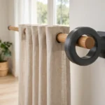 A bright home living room scene, a matte dark-gray 3D printed ring adapter fitting a wooden curtain rod into a wall bracket