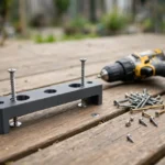A bright outdoor scene on a wooden deck, a matte charcoal-gray 3D printed deck screw spacing jig laid across a deck board with two screws driven