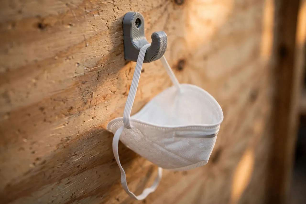 Close-up of a small 3D printed wall hook holding a dust mask by its elastic strap, the mask cup hanging clear of the wall