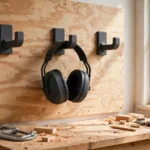 A bright home workshop wall with ear muffs hanging from matte charcoal-gray 3D printed wall hooks on a plywood panel beside a workbench, ear cups