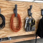 A tidy garage wall scene, several matte charcoal-gray 3D printed hooks screwed to a plywood wall holding coiled extension cords
