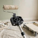 A bright home room mid-painting, a matte dark-gray 3D printed adapter joining a paint roller frame to a long extension pole