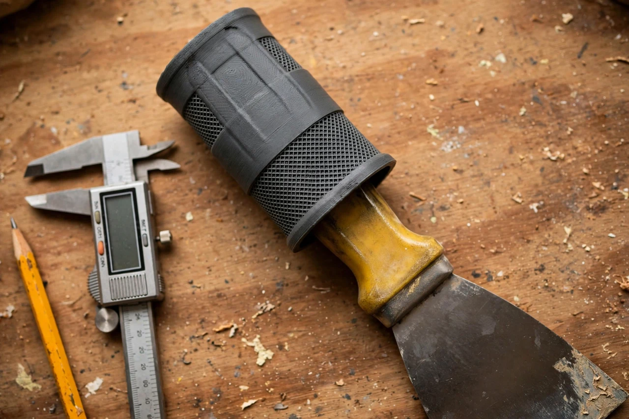 Close-up top-down view of a matte charcoal-gray 3D printed putty knife handle grip sleeve being test-fitted against a putty knife handle that slips in the hand on a workbench, calipers and a pencil nearby, warm workshop lighting