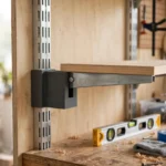 A bright home workshop scene, a matte dark-gray 3D printed adapter joining a metal shelf bracket to a slotted wall upright