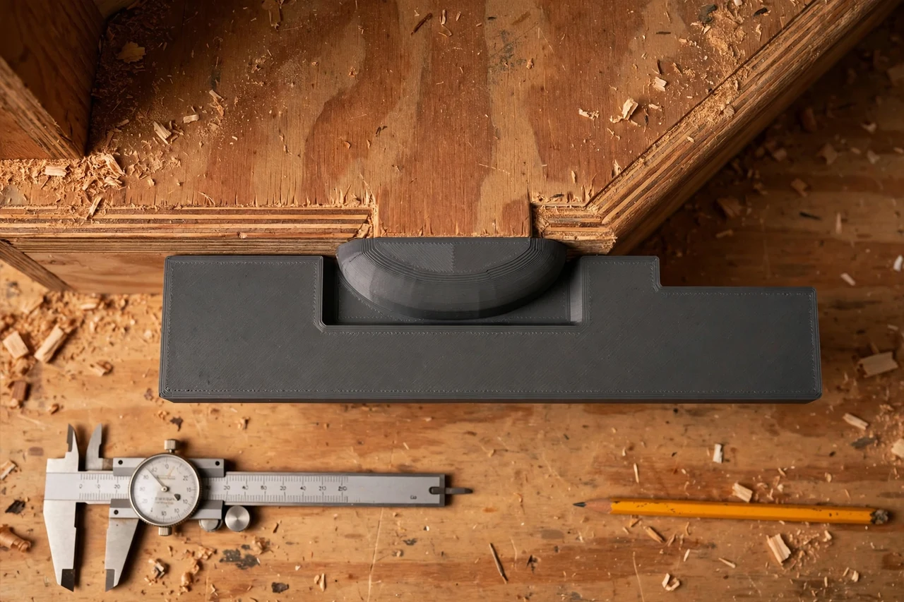 Close-up top-down view of a matte charcoal-gray 3D printed stair tread nosing alignment gauge being test-fitted against stair treads and nosing edges on a workbench, calipers and a pencil nearby, warm workshop lighting