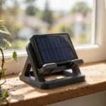 A bright window scene with a small sealed solar charger resting in a matte charcoal-gray 3D printed window ledge mount on an inside windowsill