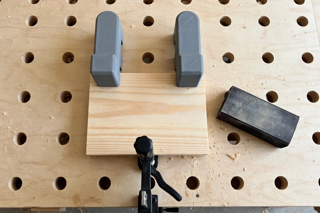 Top-down view of a small wooden panel held on a workbench between two 3D printed bench dogs at one end and a low-profile hold-down clamp