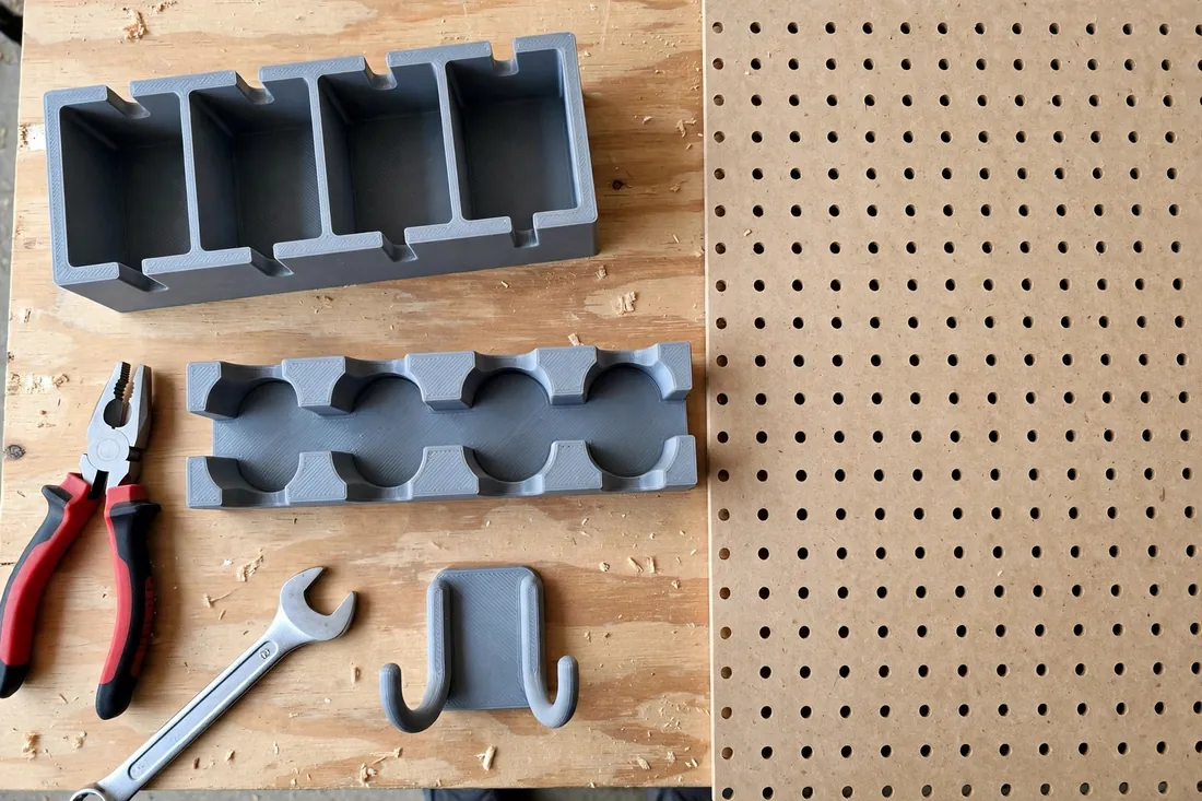 Top-down detail of three 3D printed pegboard holders on a workbench beside a section of pegboard — a pliers rack with divided slots